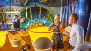 Eine Bar mit Bartender, einer Frau mit einem Getränk und einem Mann in weißer Hemd. Gelbe Stühle und ein Pool sind im Hintergrund zu sehen.