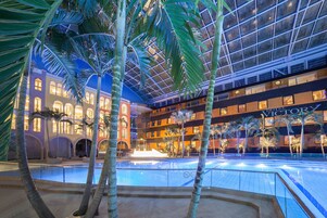 Una piscina interior con palmeras, techo de cristal y el hotel HOTEL VICTOR al fondo.