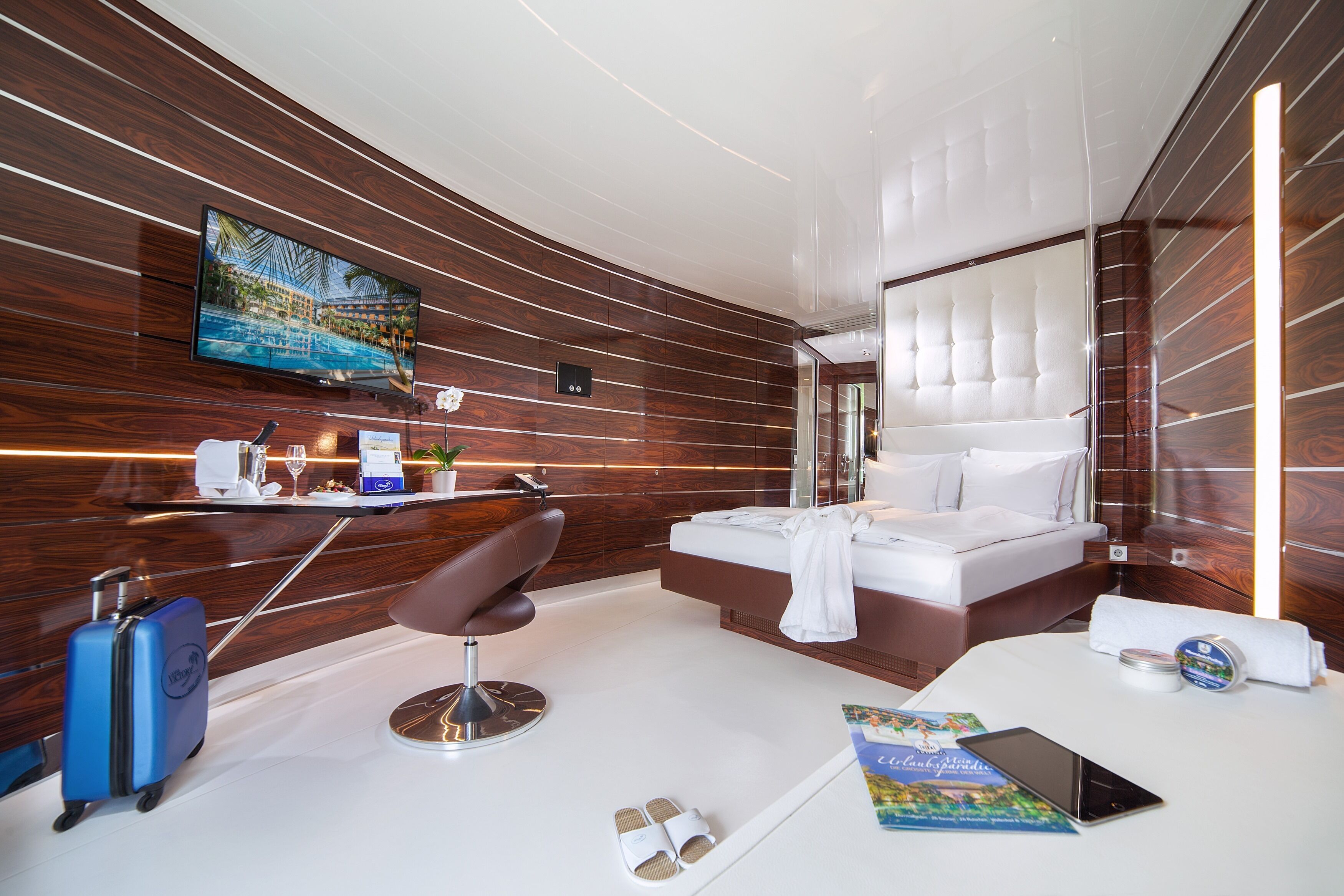 A modern hotel room with a large bed, a sitting area with a chair, a desk with a flat-screen TV, a suitcase, and a book on the floor.