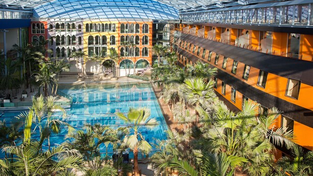 An indoor swimming pool with a glass ceiling, surrounded by palm trees and a multi-story building with arched windows. - Hotel Victory Therme Erding (Erding)