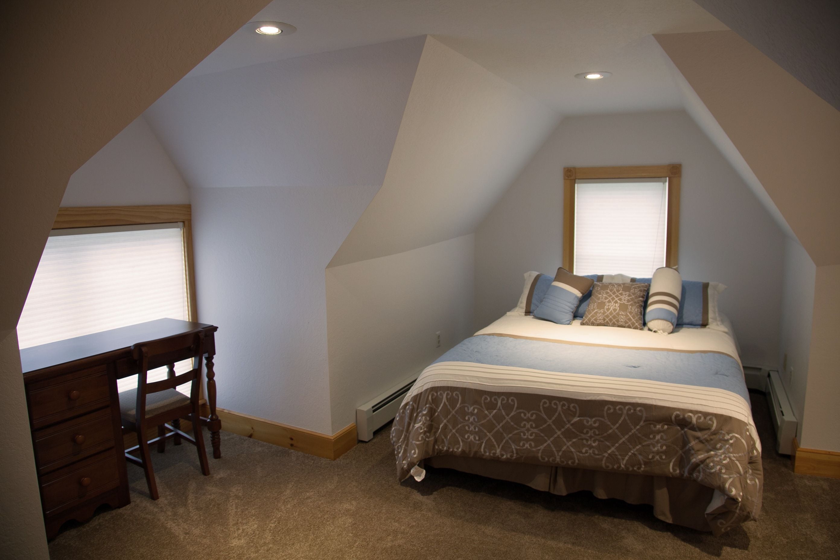 Een slaapkamer met een schuine kap, een bed met beddengoed met een patroon, een houten bureau en een raam met jaloezieën.