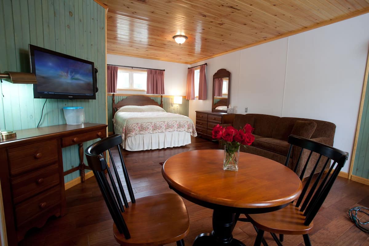 Une pièce chaleureuse avec un plafond en bois, un lit recouvert d’une courtepointe à motifs floraux, un canapé marron, un bureau en bois avec une télévision et une table ronde avec des chaises.