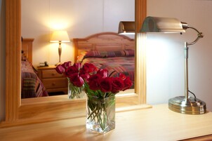 Un espejo refleja una cama, una lámpara de noche y un florero con rosas rojas sobre un tocador de madera.