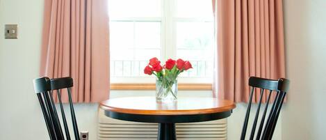 A dining area with a round table, two chairs, a vase with red flowers, and a window with pink curtains.