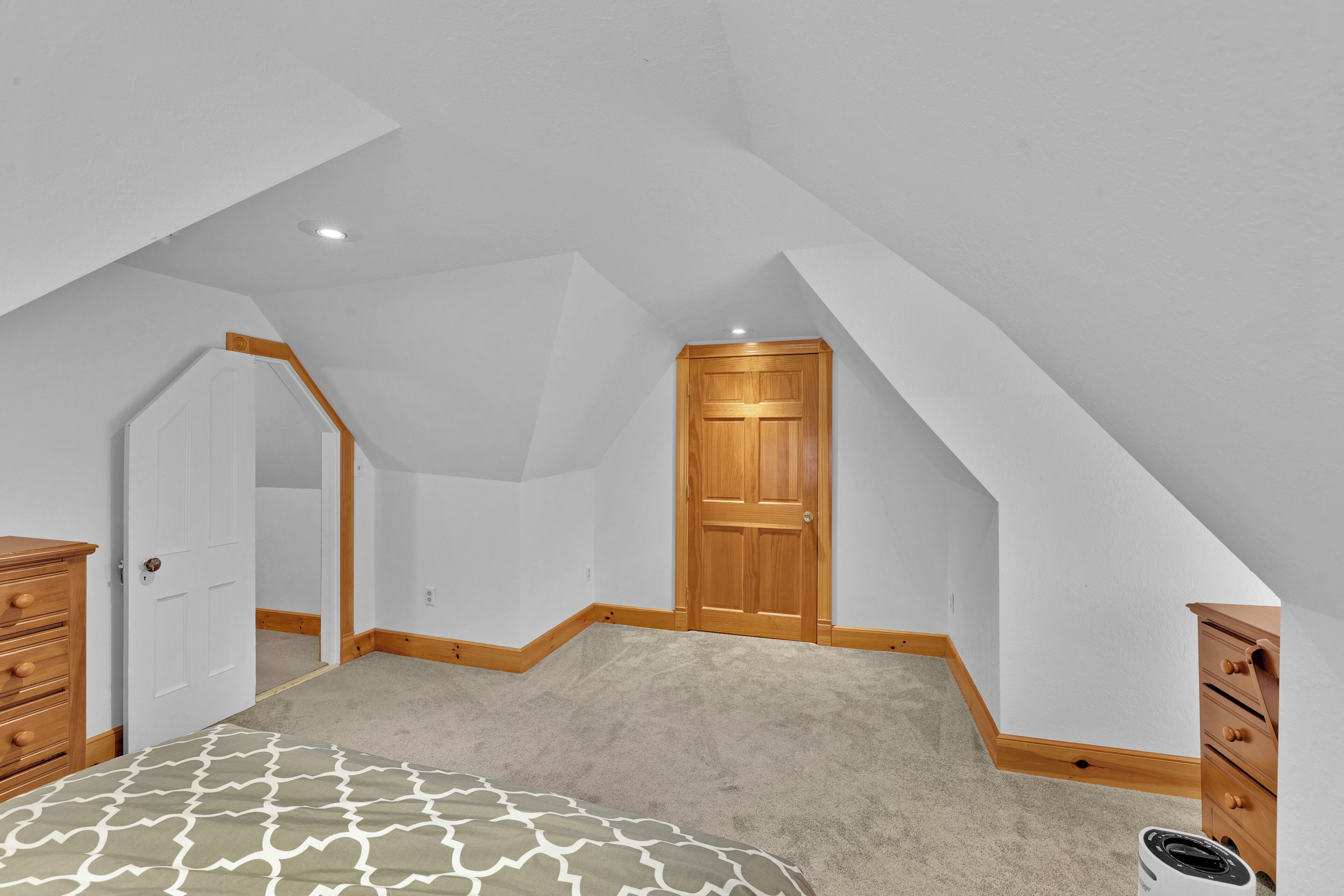 A bedroom with a sloped ceiling, a wooden door, a dresser, and a patterned bedspread.