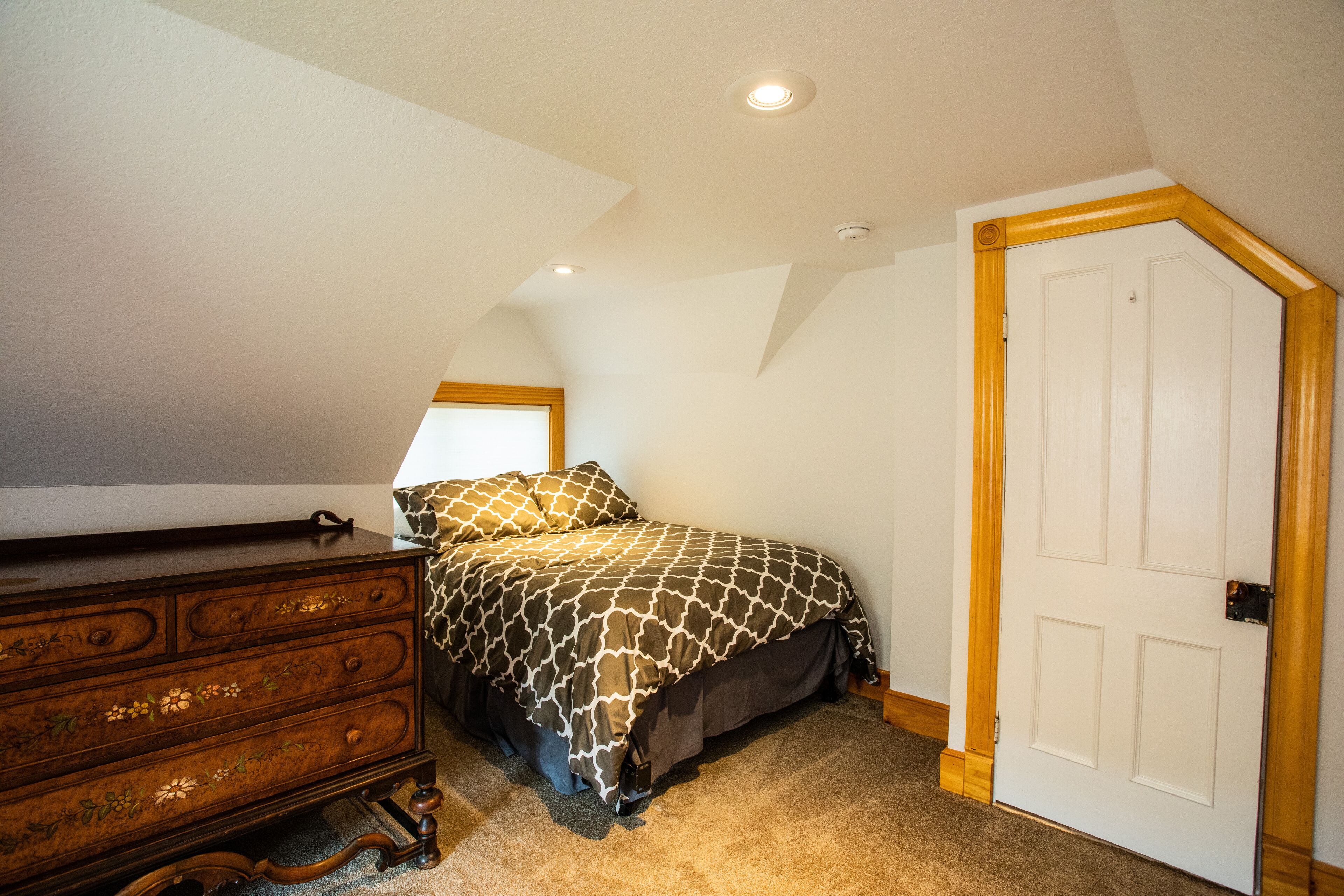A bedroom with a sloped ceiling, a dresser with floral carvings, a patterned bedspread, and a white door with a wooden frame.