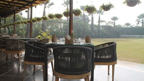 Restaurante ao ar livre com cadeiras de vime, vasos suspensos e vista para o gramado.