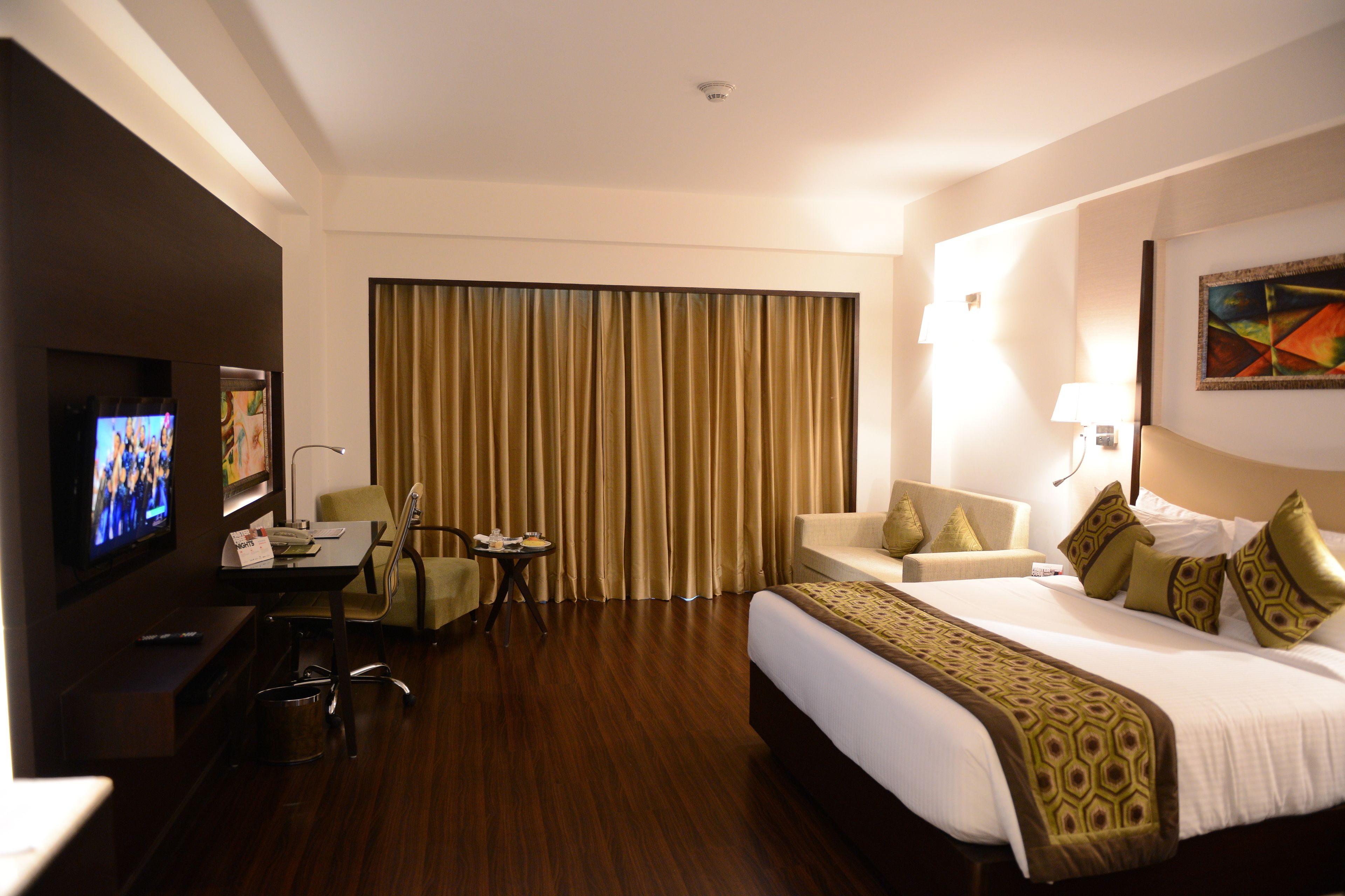 Une chambre d’hôtel avec un grand lit, un bureau avec une chaise, une télévision et une grande fenêtre avec des rideaux.
