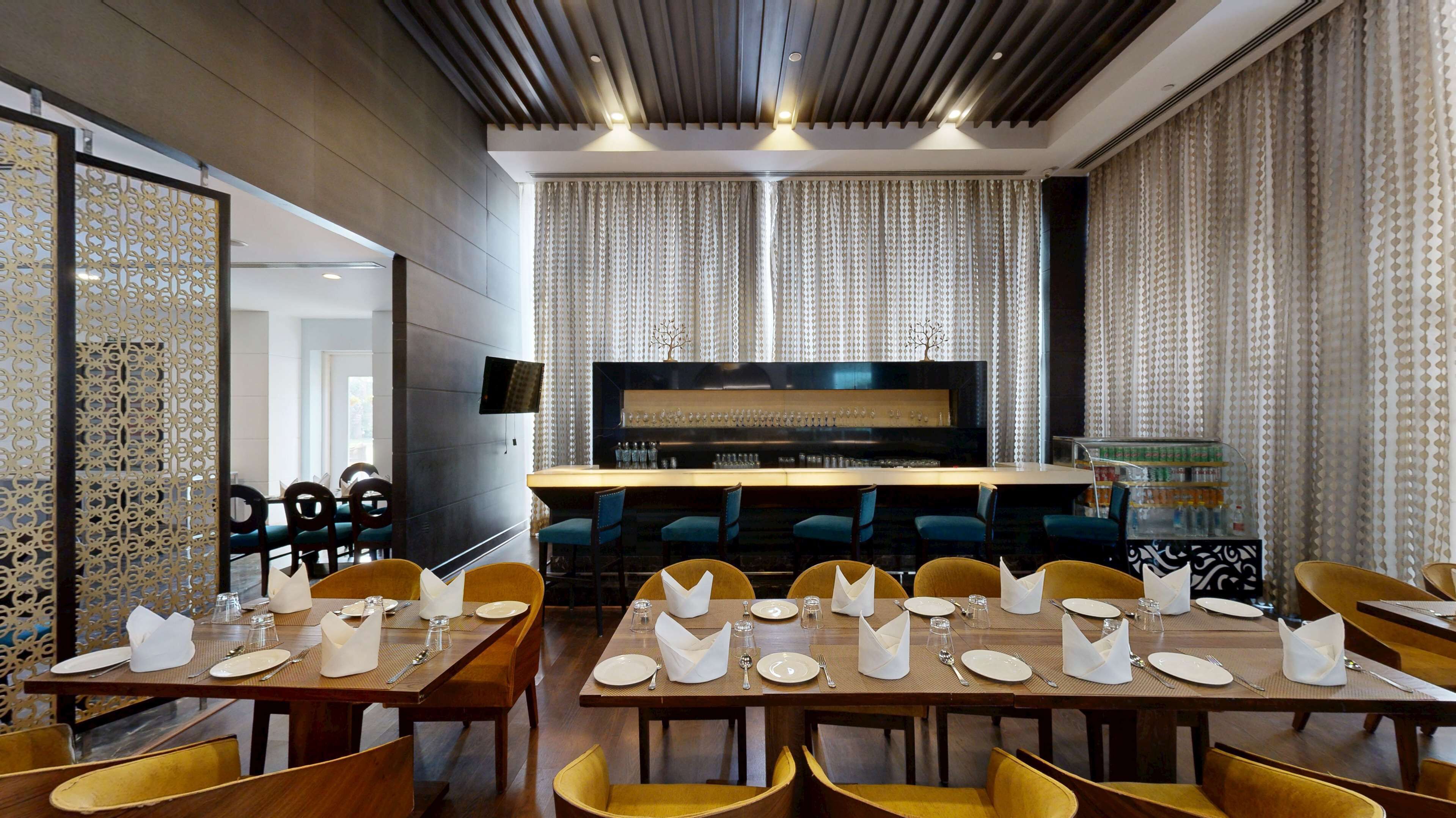 A restaurant with a modern interior, featuring a long counter with blue chairs, a television mounted on the wall, and a decorative screen with a tree design.