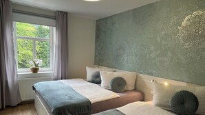 A modern bedroom with two beds, a large window with curtains, a wall-mounted light fixture, and a decorative wall piece. - Penzion Jasmín (Liberec)
