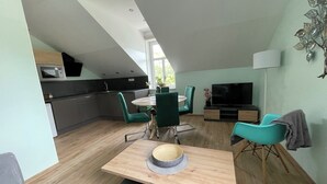 A modern kitchen with a microwave and cabinets, a dining area with a wooden table and green chairs, and a living area with a TV and a decorative plant. - Penzion Jasmín (Liberec)