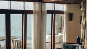 A room with a large window overlooking the sea, a wooden ceiling, and a view of a white chair and table outside. - SONG SAA PRIVATE ISLAND (Koh Ouen)