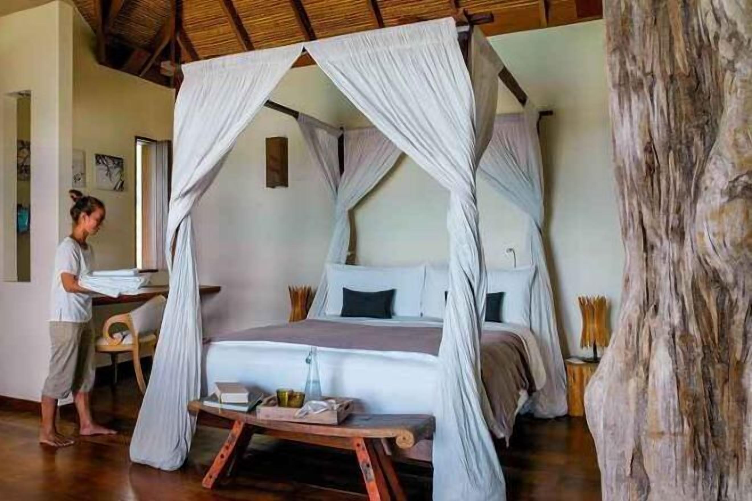 Una cama con dosel y cortinas blancas, una mesita de noche de madera y un tronco de árbol rústico en la habitación.