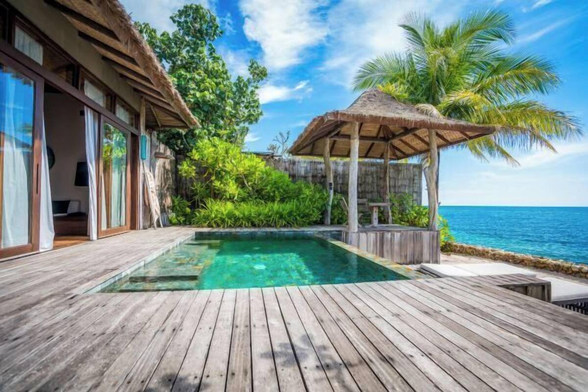 Una terraza de madera con piscina, una estructura de techo de paja y vistas al mar.