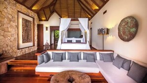 A bedroom with a stone wall, a bed with white bedding, a wooden ceiling, and a sofa with grey cushions. - SONG SAA PRIVATE ISLAND (Koh Ouen)