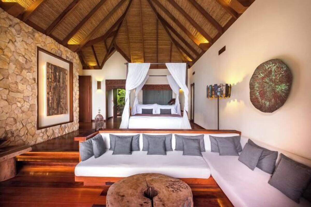 Un dormitorio con pared de piedra, cama con ropa de cama blanca, techo de madera y sofá con cojines grises.