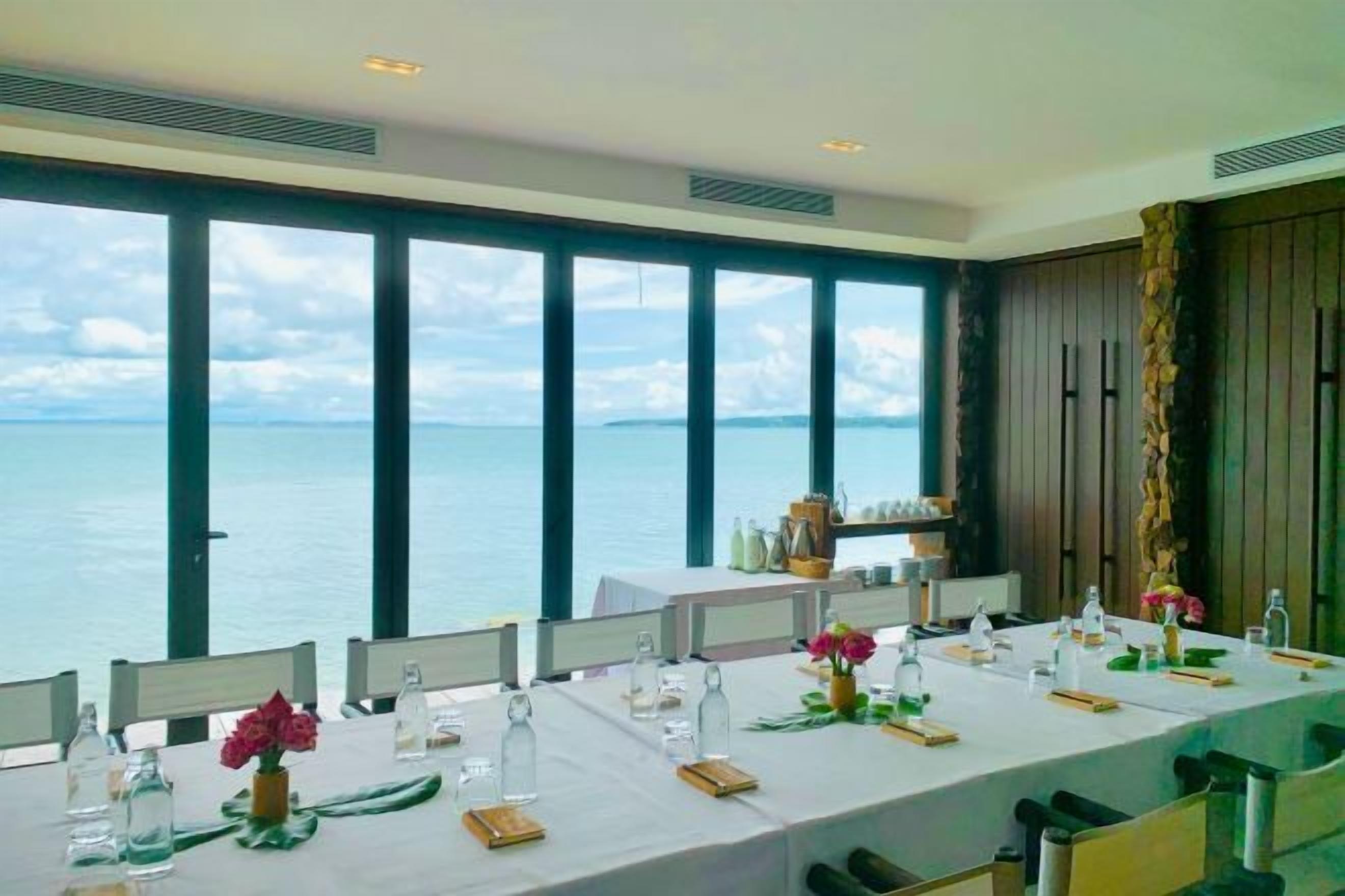 A dining area with a view of the sea, large windows, and a long table set for a meal.