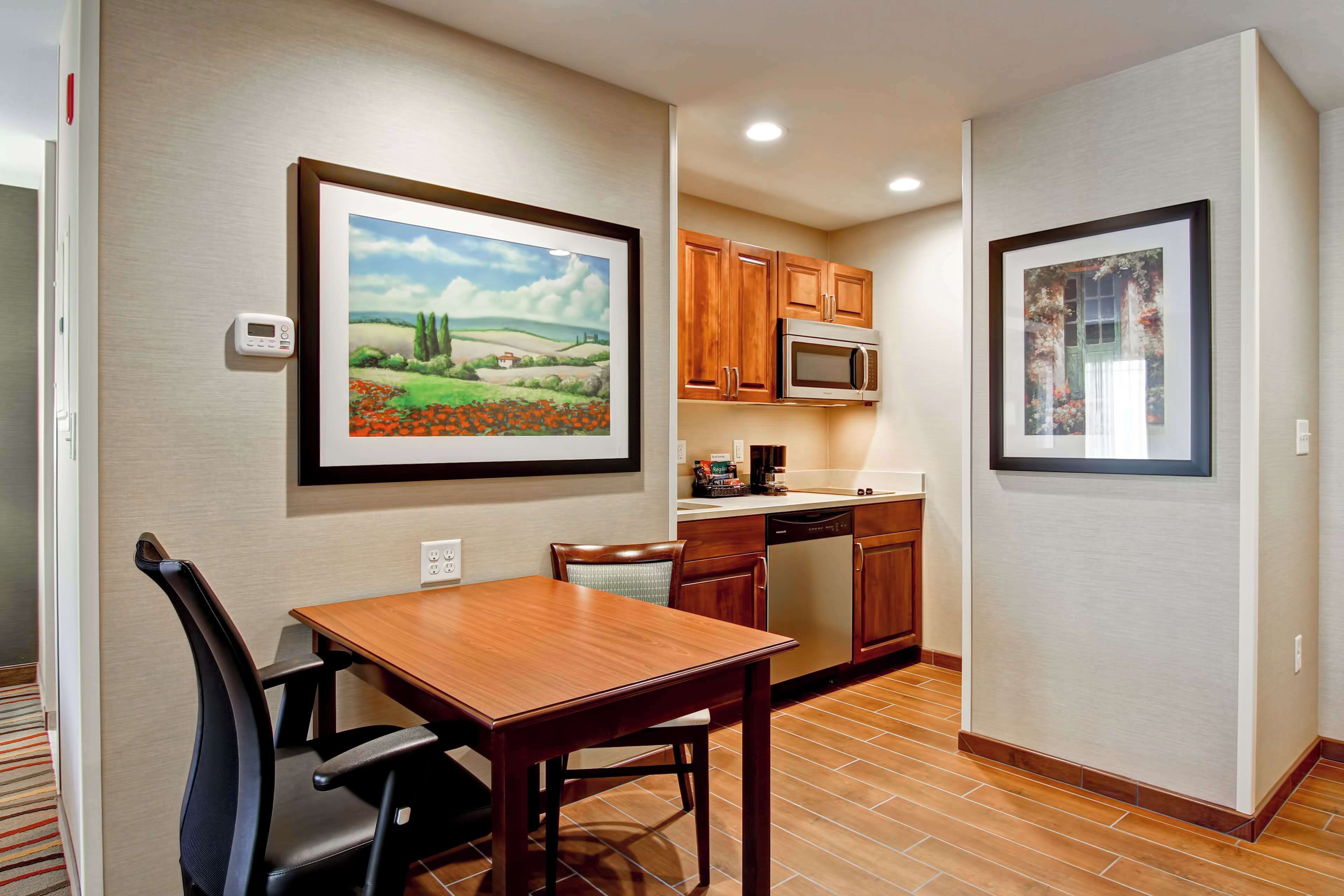 A compact kitchen with wooden cabinets, a microwave, and a dishwasher. There is a dining table with chairs, and a framed painting on the wall.