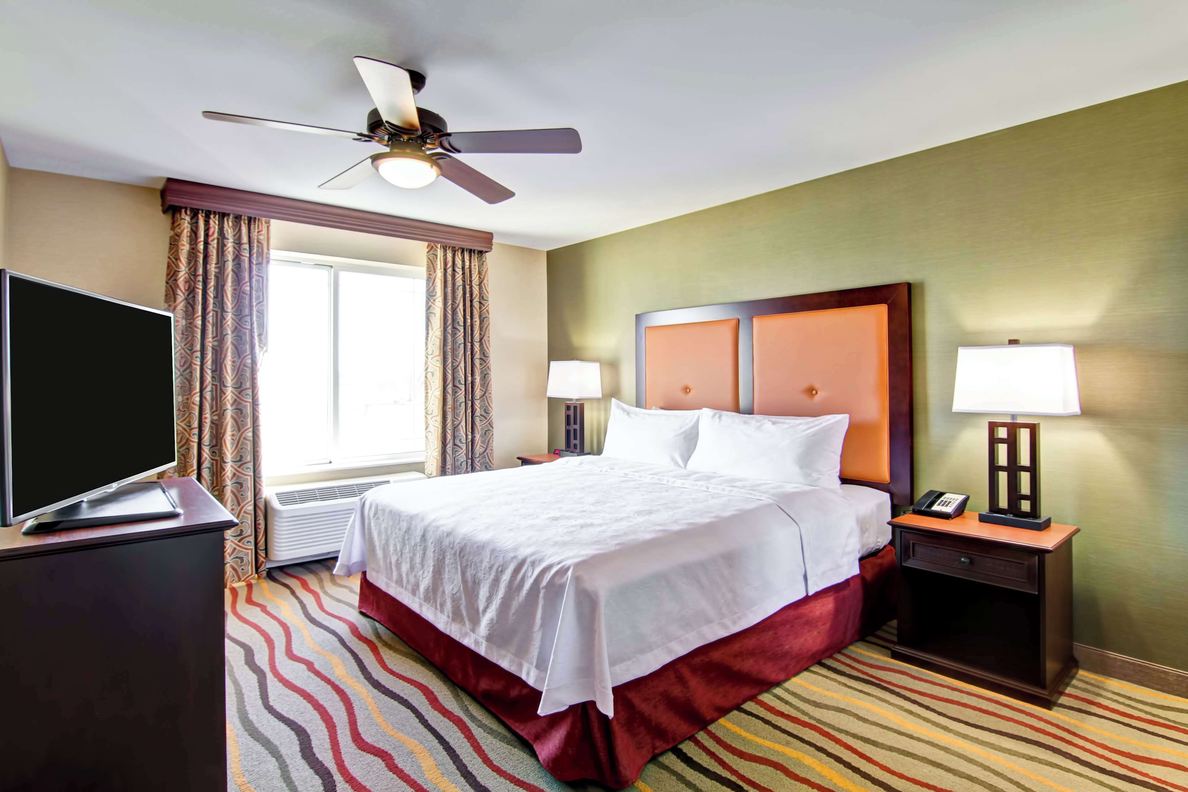 A hotel room with a bed, a TV, a desk, a chair, a lamp, and a ceiling fan.