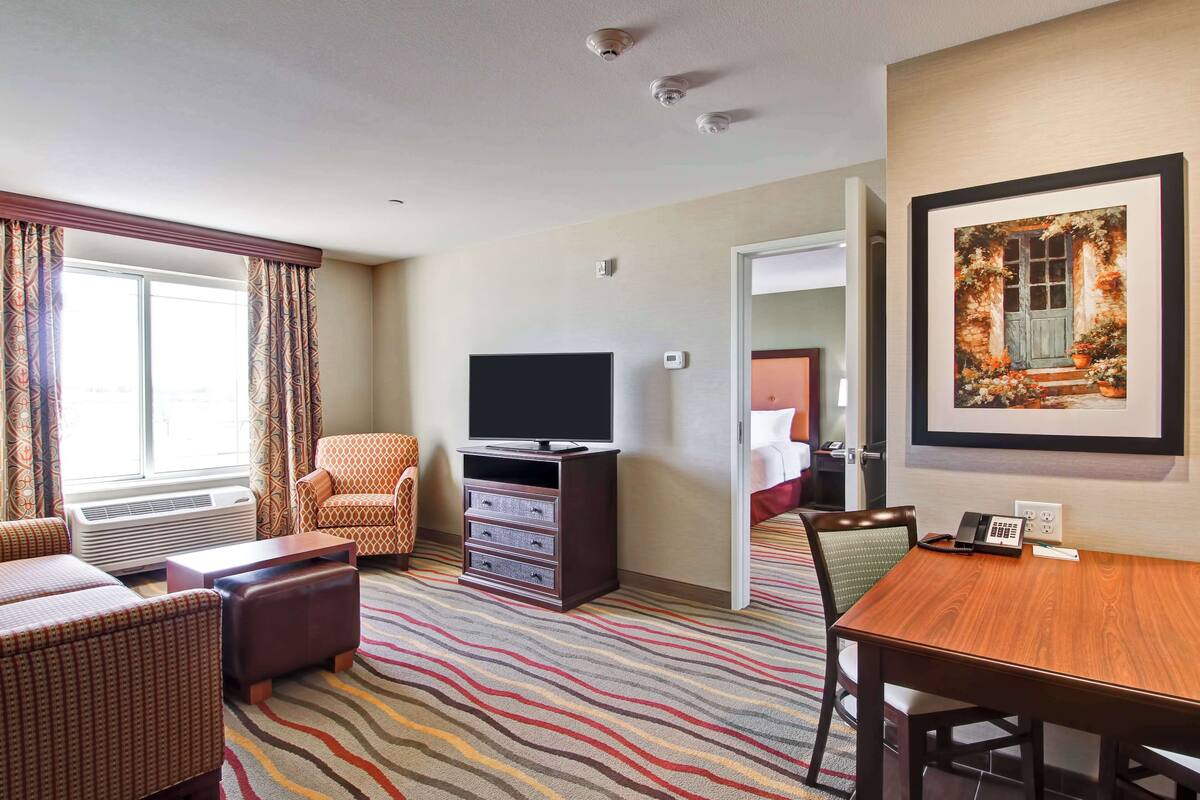 Une chambre d’hôtel dotée d’une grande fenêtre, d’un téléviseur à écran plat, d’une table à manger, d’un canapé, d’un fauteuil et d’un tapis à motifs.