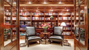 Una biblioteca con sillones de cuero, una mesa redonda con un florero y estanterías llenas de libros.