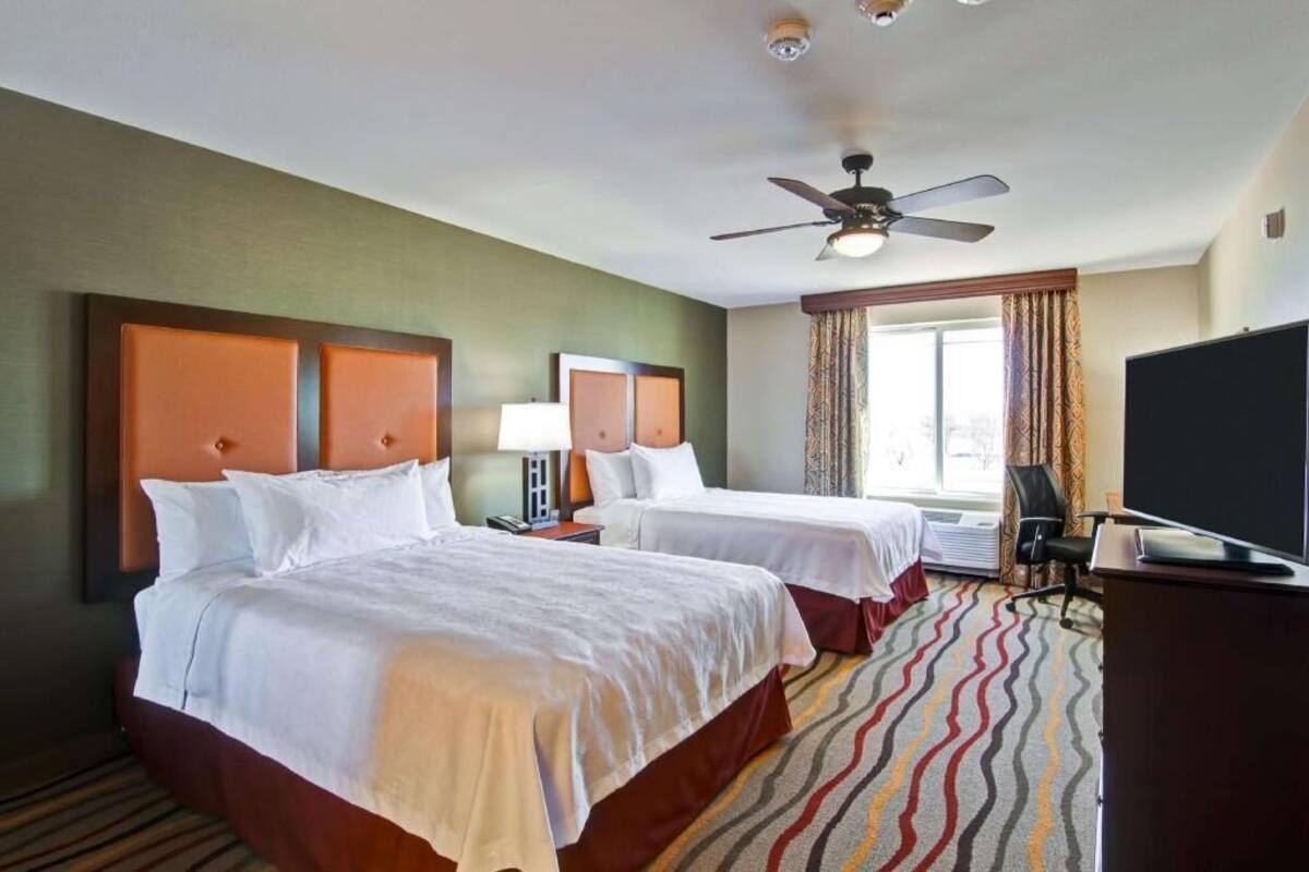 Une chambre d’hôtel avec deux lits, un ventilateur de plafond, une télévision et une fenêtre avec des rideaux.