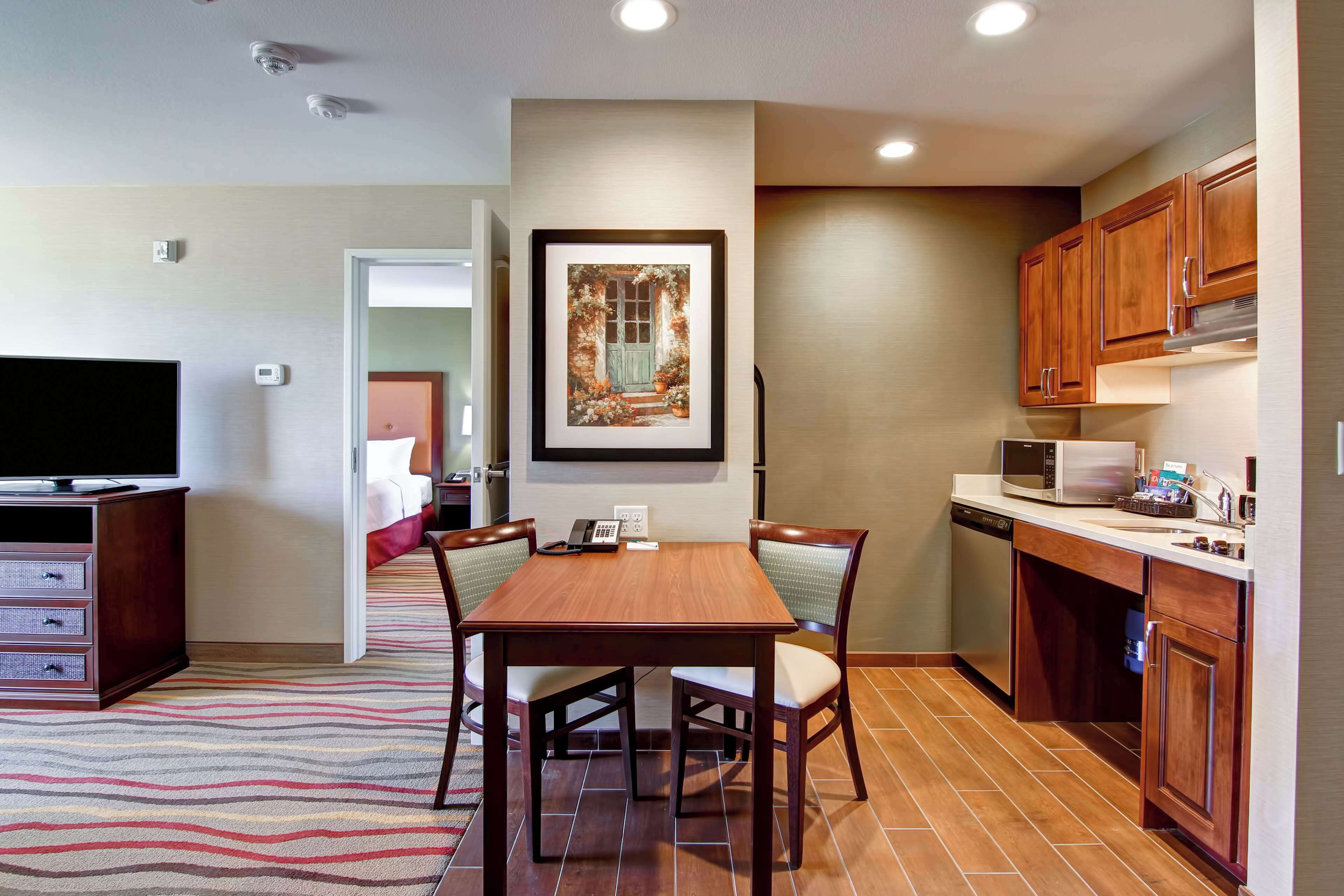 A modern kitchen with wooden cabinets, a microwave, and a dishwasher. There is a dining table with chairs and a framed picture on the wall.