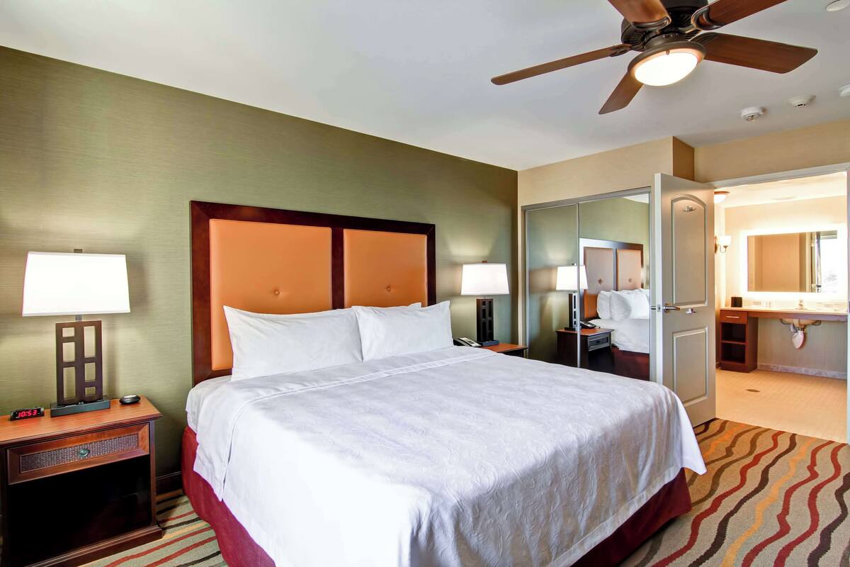 Une chambre d’hôtel comprenant un lit, des lampes de chevet, une table de chevet, une horloge, un ventilateur de plafond et une salle de bain avec un miroir et un lavabo.