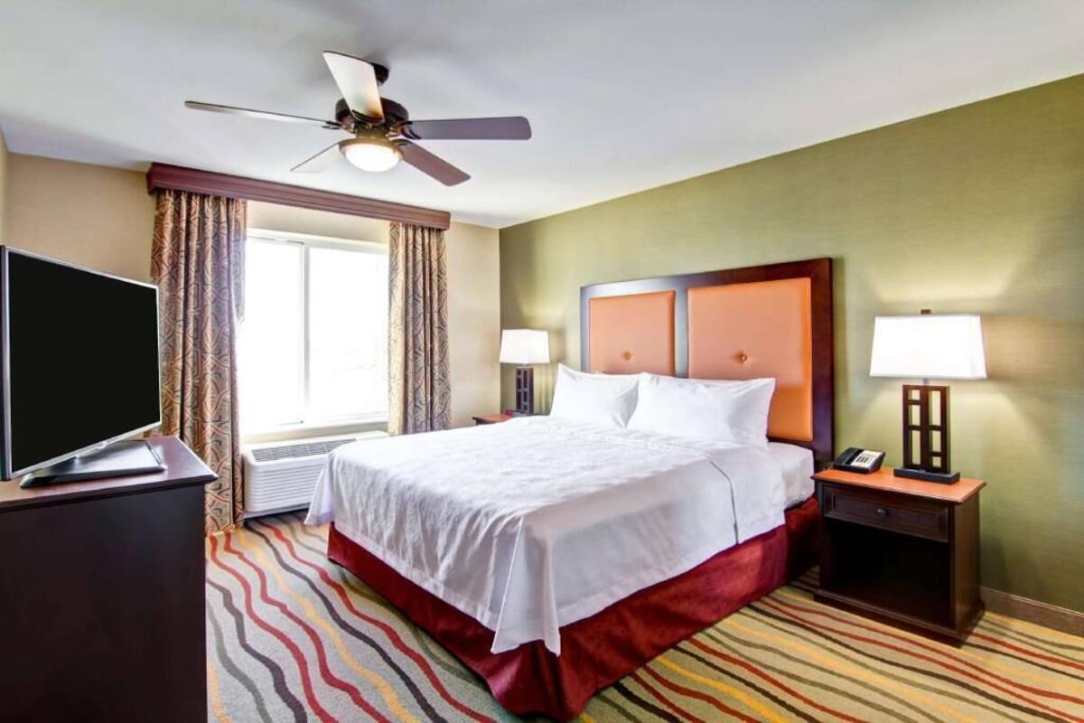 Une chambre d’hôtel comprenant un lit, une télévision, un ventilateur de plafond, une table de chevet avec une lampe et une fenêtre avec des rideaux.