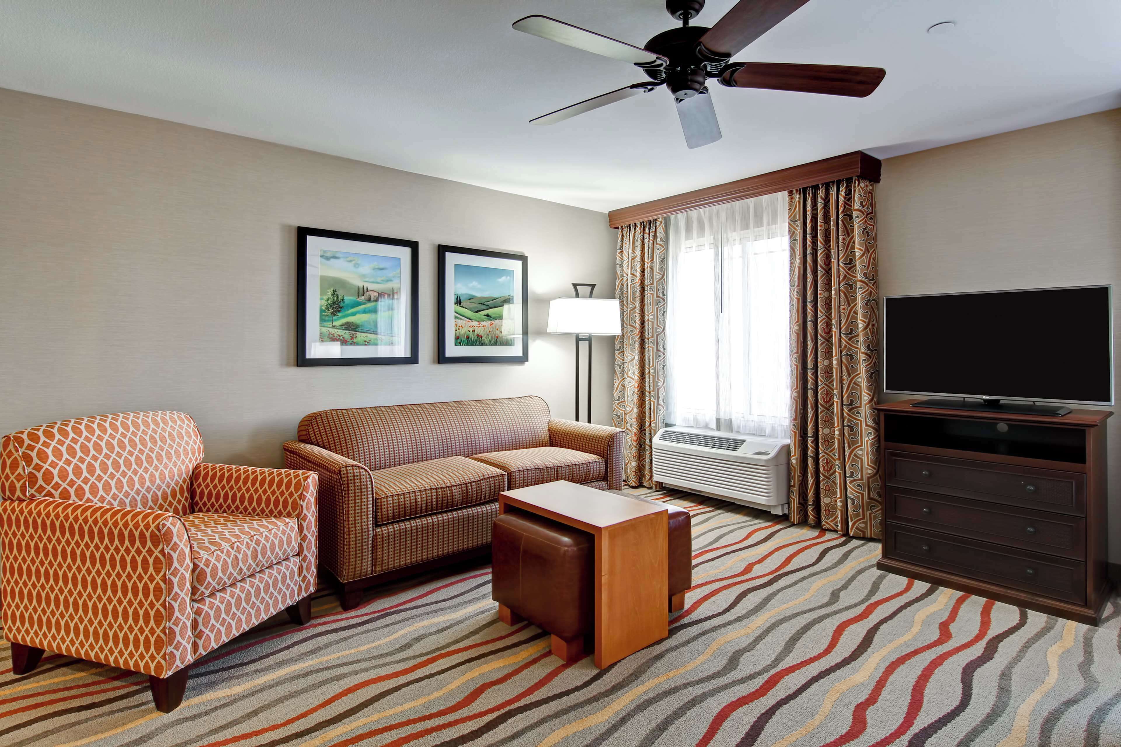 A hotel room with a sofa, armchair, small table, TV stand, ceiling fan, and two framed pictures on the wall.