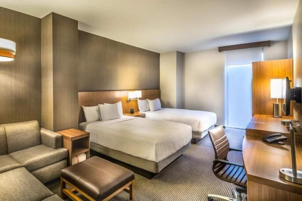 A hotel room with a sofa, two beds, a desk, a chair, and a lamp.