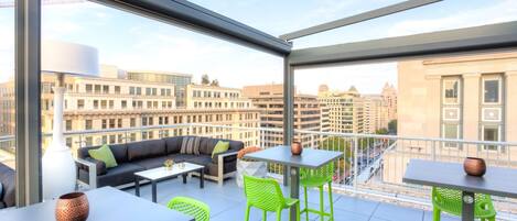 A modern outdoor balcony with a sofa, chairs, and tables, offering a city view.