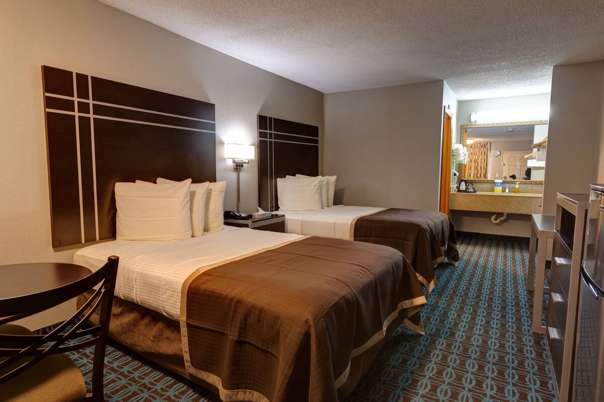 A hotel room with two beds, a desk, a chair, a small table, a mirror, and a kitchenette.