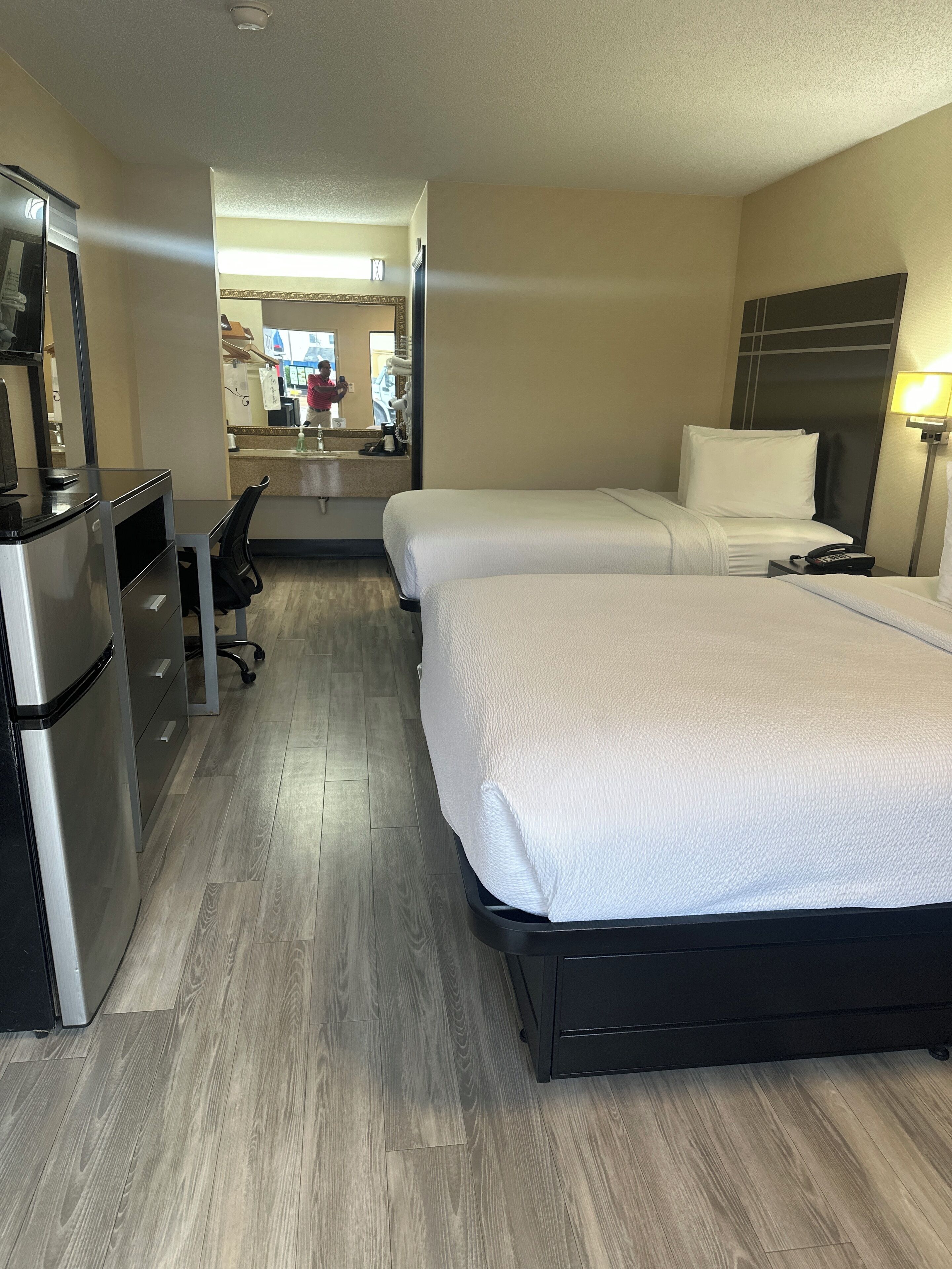A hotel room with two beds, a desk, a refrigerator, and a mirror.