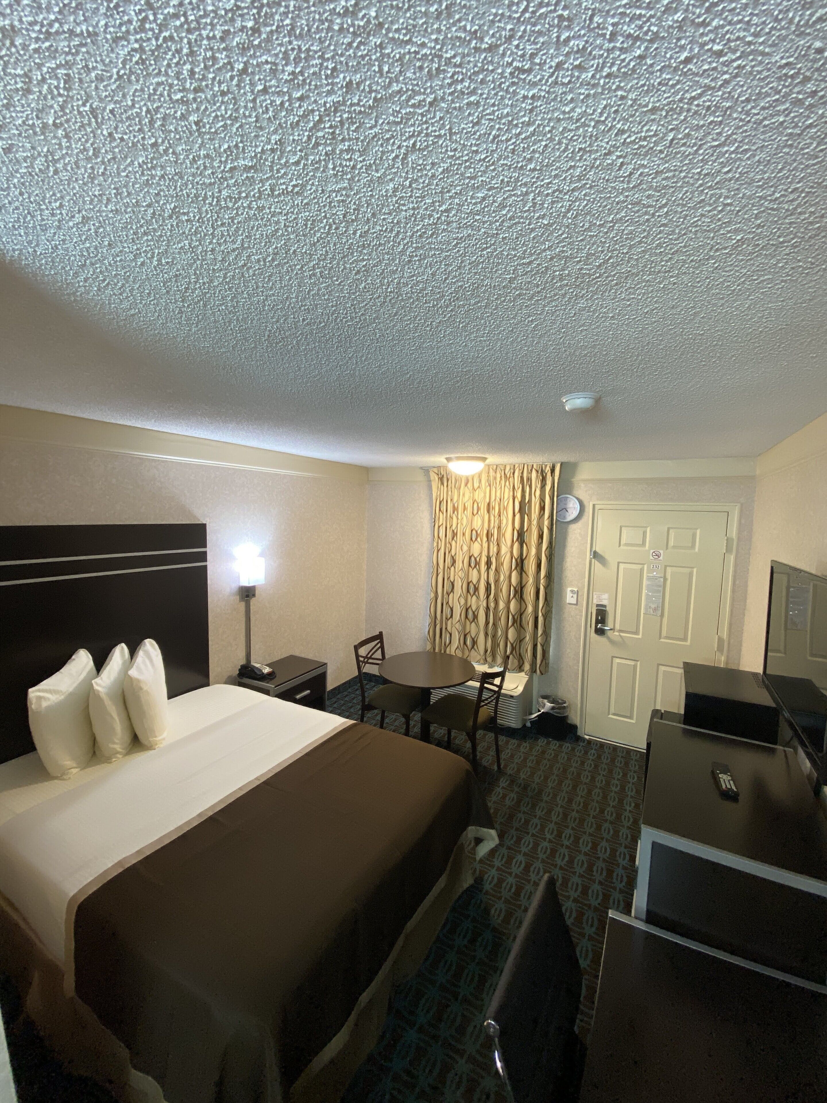 A hotel room with a bed, bedside table, lamp, desk, chair, and a door with a clock.