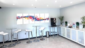 A modern bar area with white bar stools, a coffee machine, and a colorful abstract painting on the wall. - Deluxe Inn Fayetteville - I-95 Exit 49 (Fayetteville)