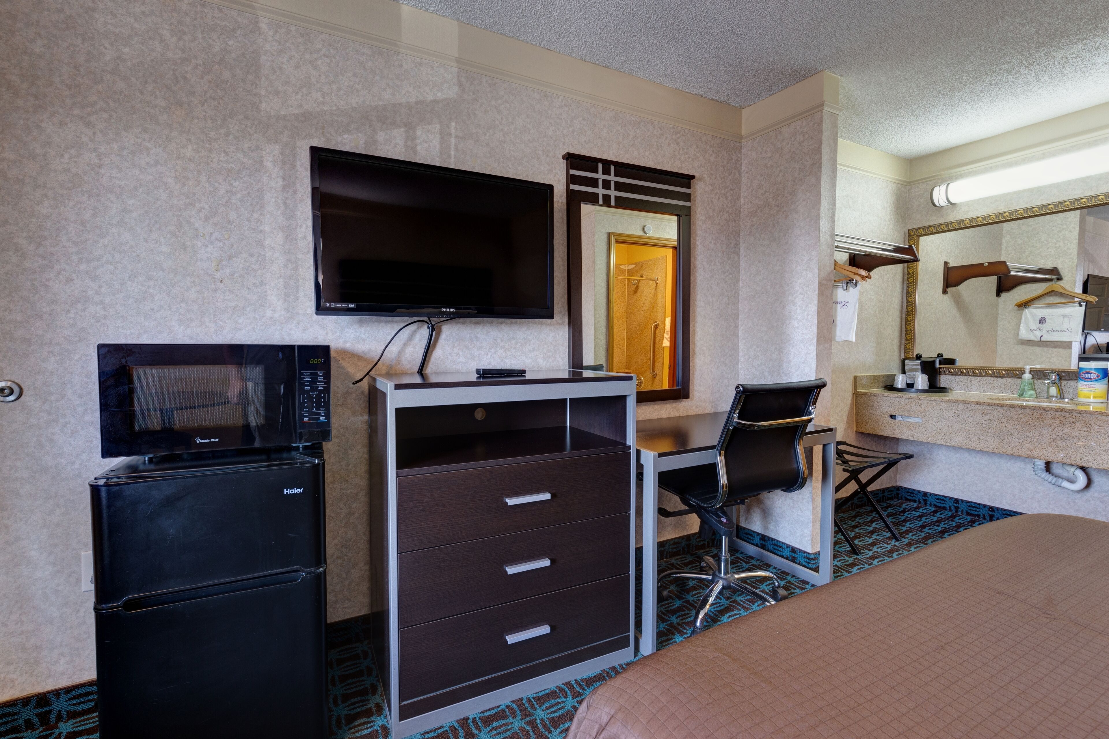 A hotel room with a bed, a desk with a chair, a television, a microwave, a refrigerator, a dresser, a bathroom with a mirror, and a door.