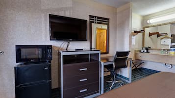 A hotel room with a bed, a desk with a chair, a television, a microwave, a refrigerator, a dresser, a bathroom with a mirror, and a door.