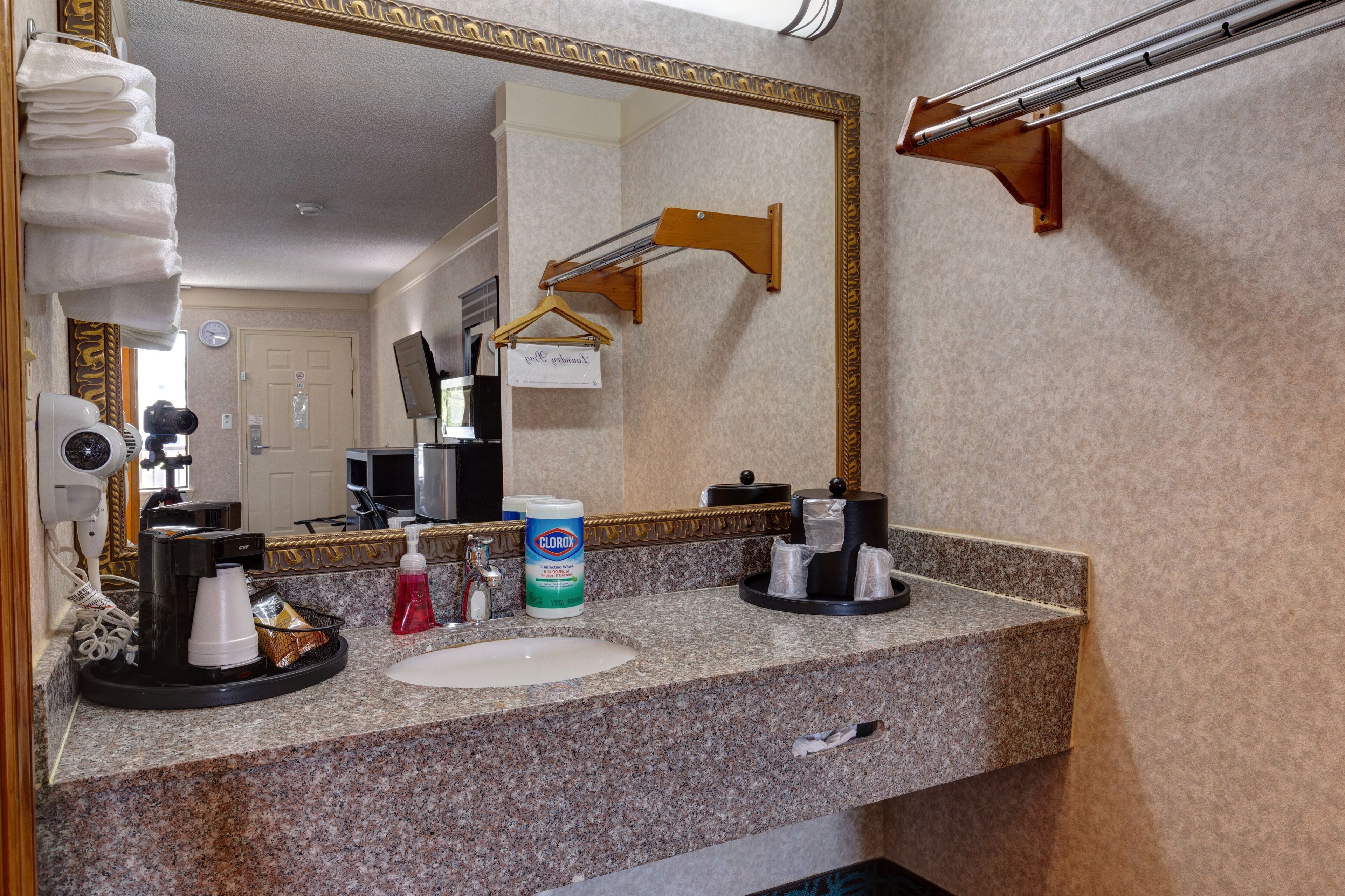 Un baño de hotel con encimera de granito, lavamanos, espejo, toallero, secador de pelo y cafetera.