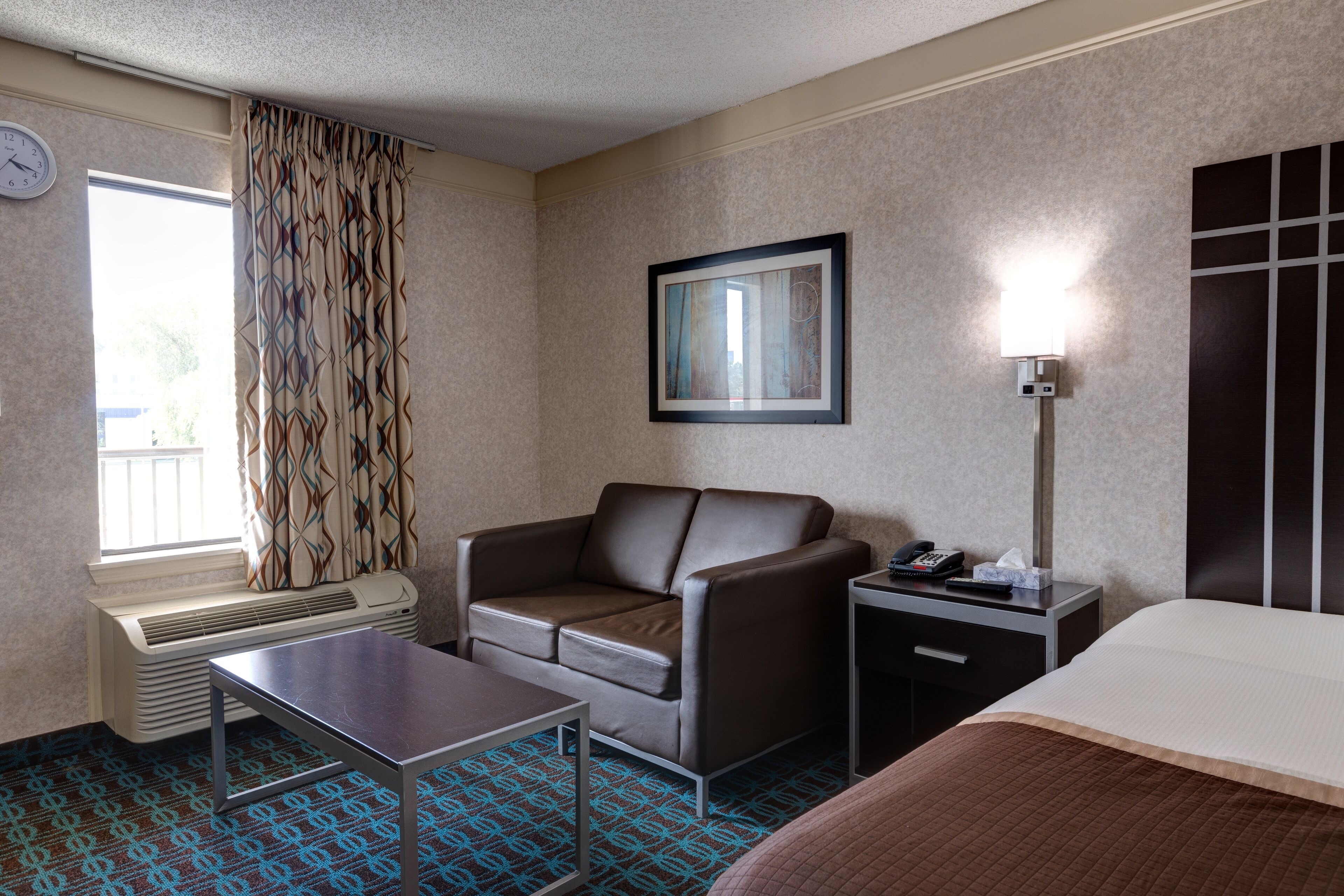 A hotel room with a brown leather sofa, a small table, a bedside table with a lamp, a bed, a window with patterned curtains, and a wall-mounted picture frame.