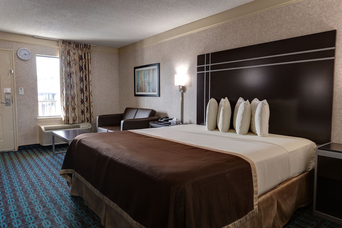 A hotel room with a large bed, a desk with a chair, a window with curtains, a wall clock, and a painting.