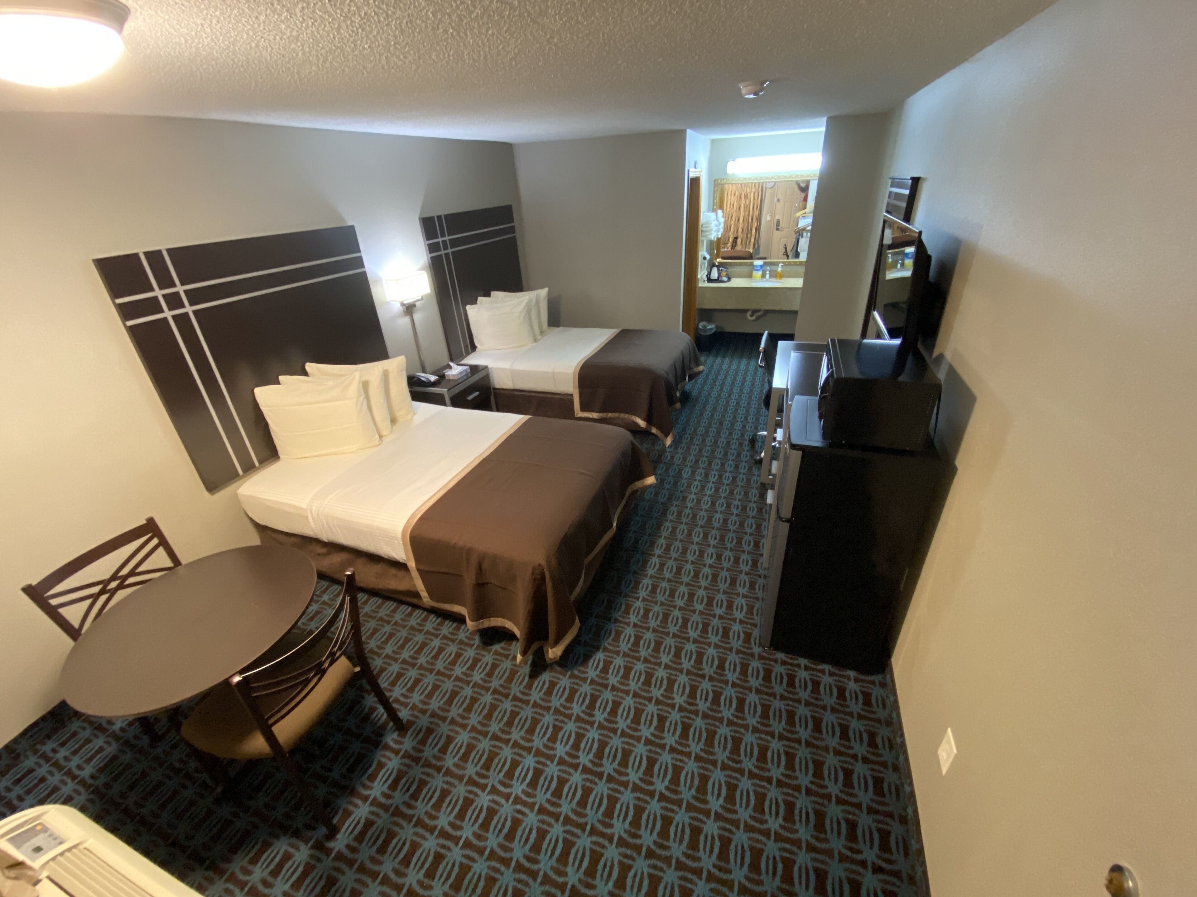 Habitación de hotel con dos camas, un escritorio, una silla, un televisor y un baño visible a través de una puerta abierta.