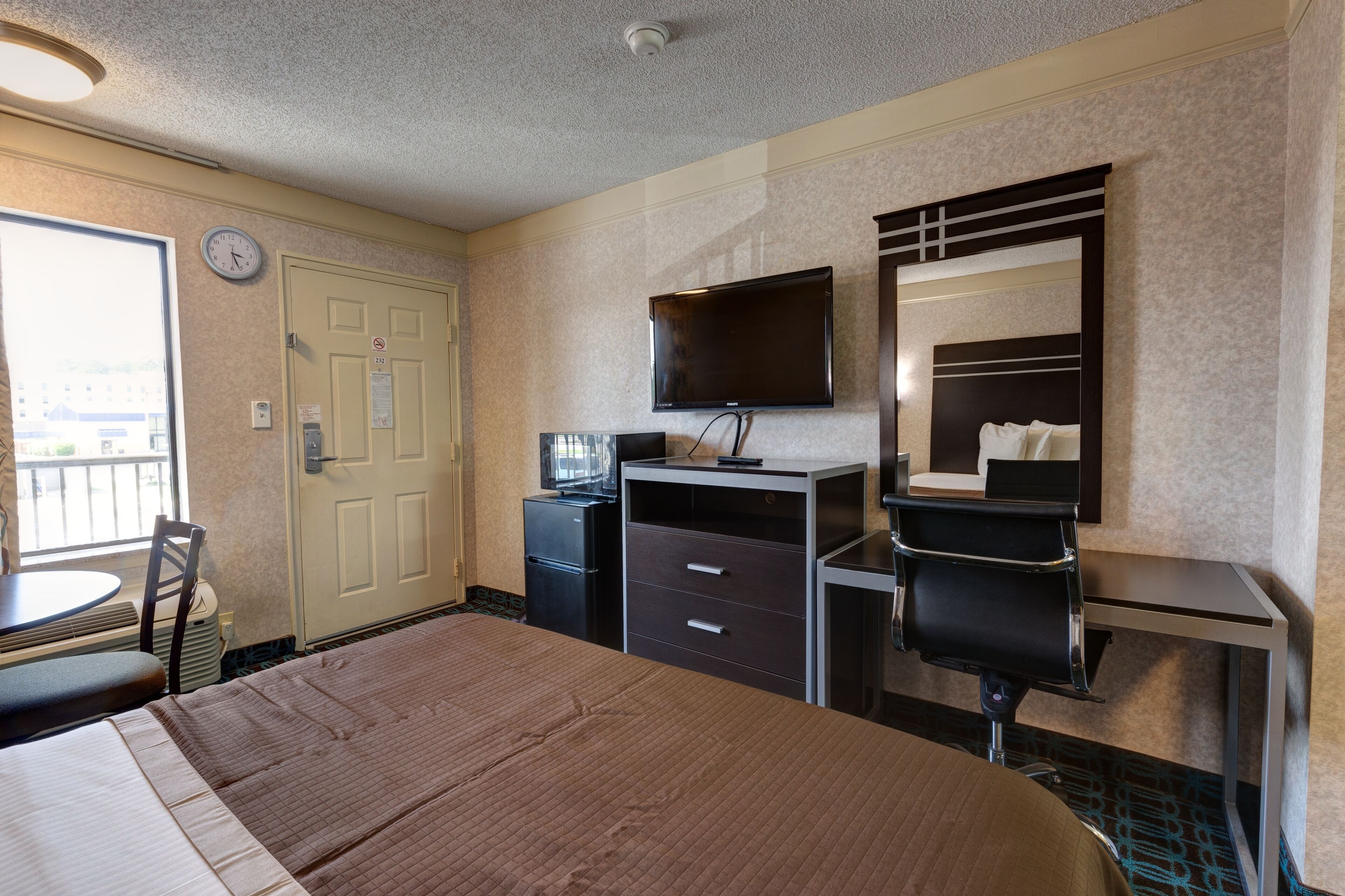 Habitación de hotel con cama, escritorio, silla, televisor, espejo, reloj y ventana.