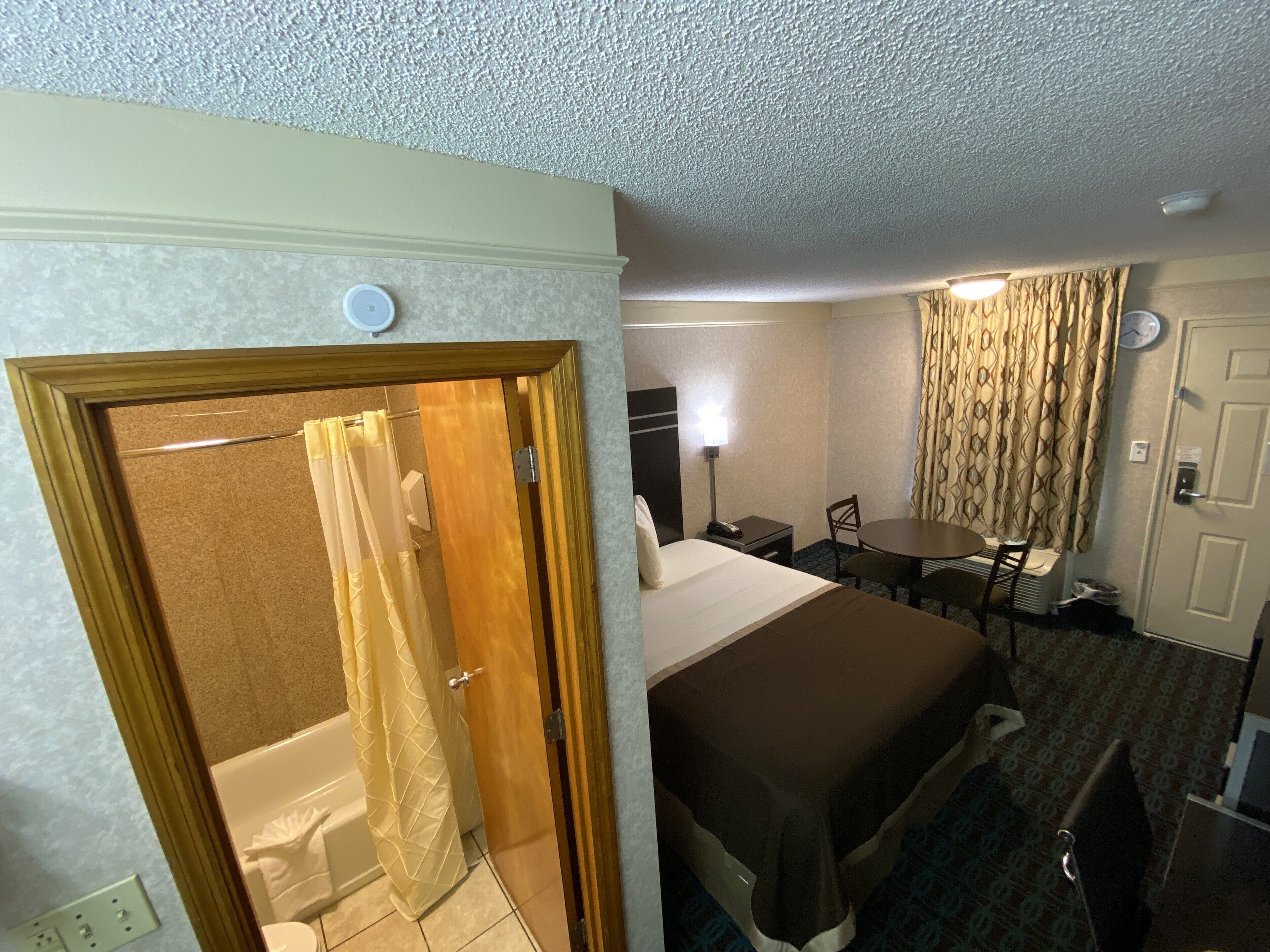 A hotel room with a bed, a table, a chair, a shower, and a toilet.