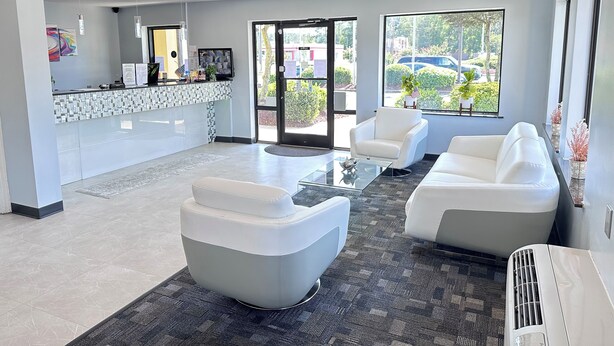 A modern reception area with a glass desk, white sofas, and a large window offering a view of the outdoors. - Deluxe Inn Fayetteville - I-95 Exit 49 (Fayetteville)