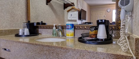 Un baño de hotel con encimera de granito, un lavabo, un espejo, un secador de pelo, una cafetera y una botella de jabón de manos.
