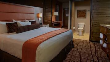 A hotel room with a large bed, bedside tables, a desk, a chair, a bathroom with a toilet, sink, and mirror.
