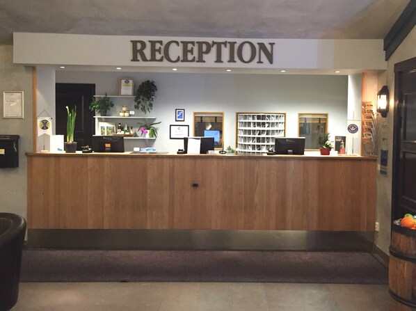 Un mostrador de recepción con un cartel que dice 'RECEPTION', una computadora, una planta y un estante con varios objetos.
