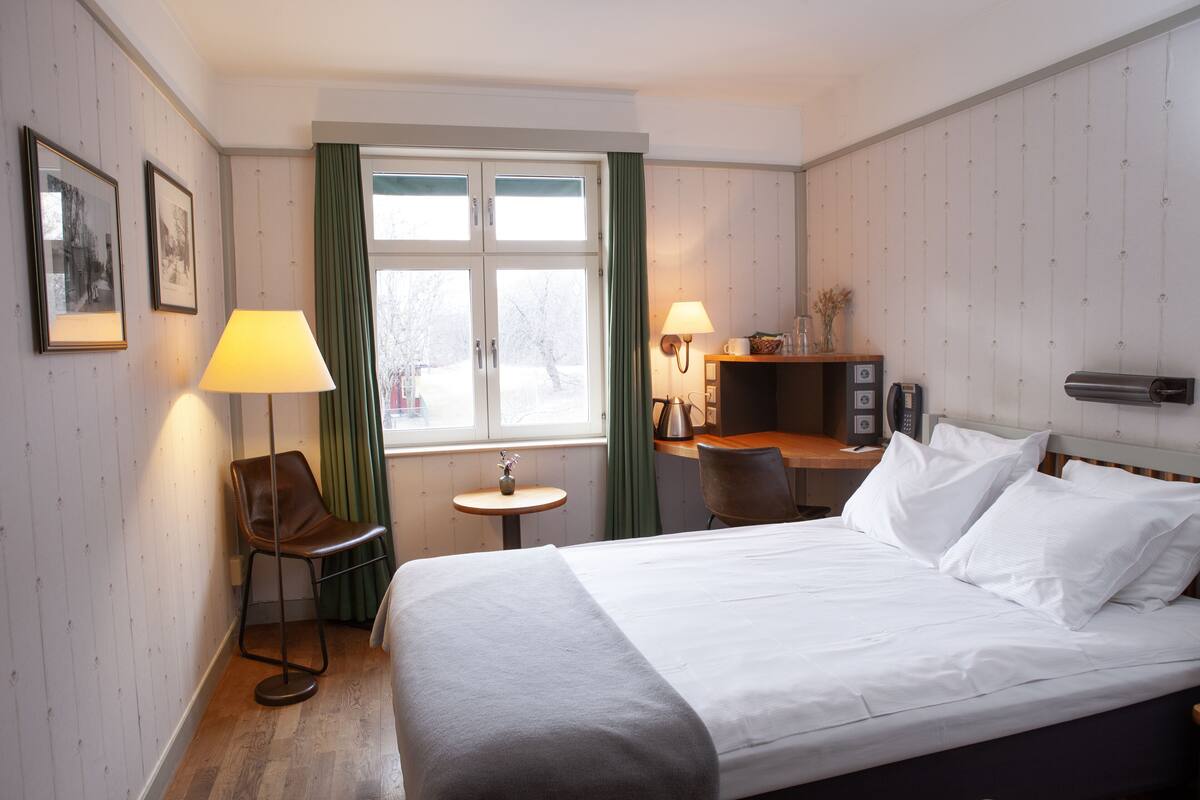 A hotel room with a bed, a desk, a chair, a lamp, and a window.