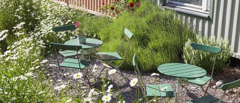 Un espace jardin aménagé avec des chaises et des tables en métal vert, entouré de fleurs blanches et roses, et un bâtiment avec une fenêtre et une porte en arrière-plan.